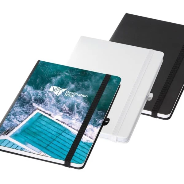 Three notebooks are shown. The first has a cover featuring an aerial view of a pool next to the ocean with Destination NSW text. The other two are plain, one white and one black. They all have elastic bands.
