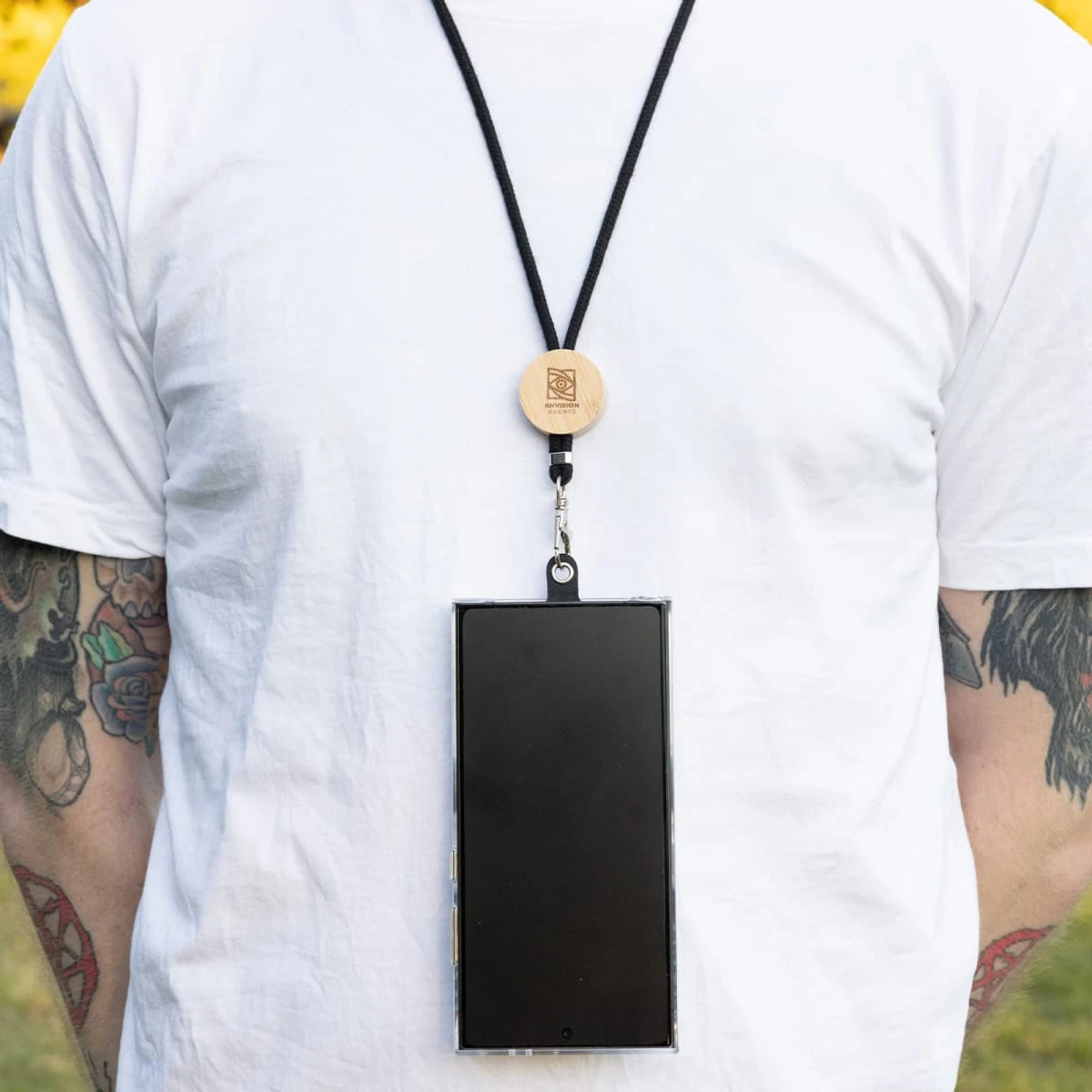 A person wearing a white T-shirt with tattoos on their arms is shown with a smartphone attached to a black lanyard around their neck. The lanyard has a wooden clasp. The background is a blurred green outdoor setting.
