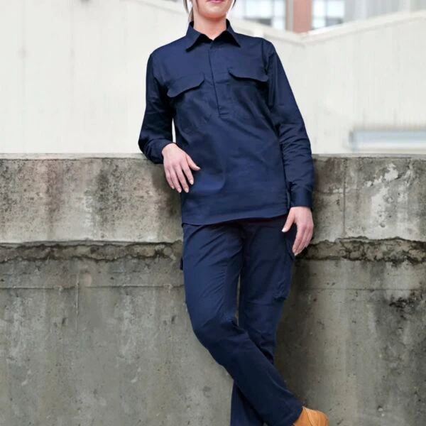 A person in navy blue workwear and tan boots leans against a concrete wall in an industrial setting. The background features buildings with large windows.