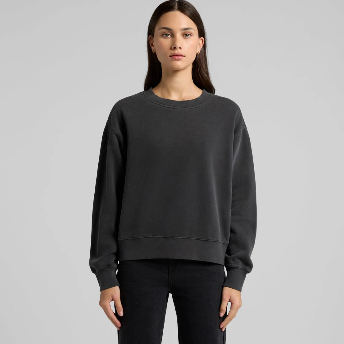 Woman in a plain black sweatshirt and black jeans standing against a gray background.
