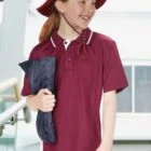 Child in maroon hat and polo shirt smiling, holding a navy jacket indoors.