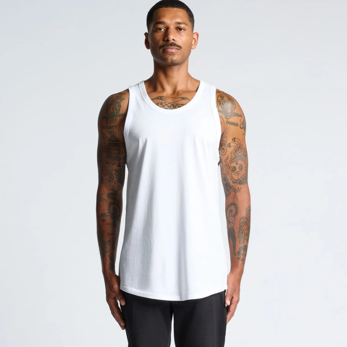Man with tattoos wearing a white tank top and black pants, standing against a plain background.