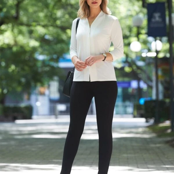 Person in a white shirt and black pants standing outdoors on a paved path with trees in the background.