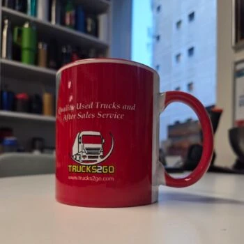 Red Trucks2Go ceramic mug on a white desk with office shelves and a window in the background.